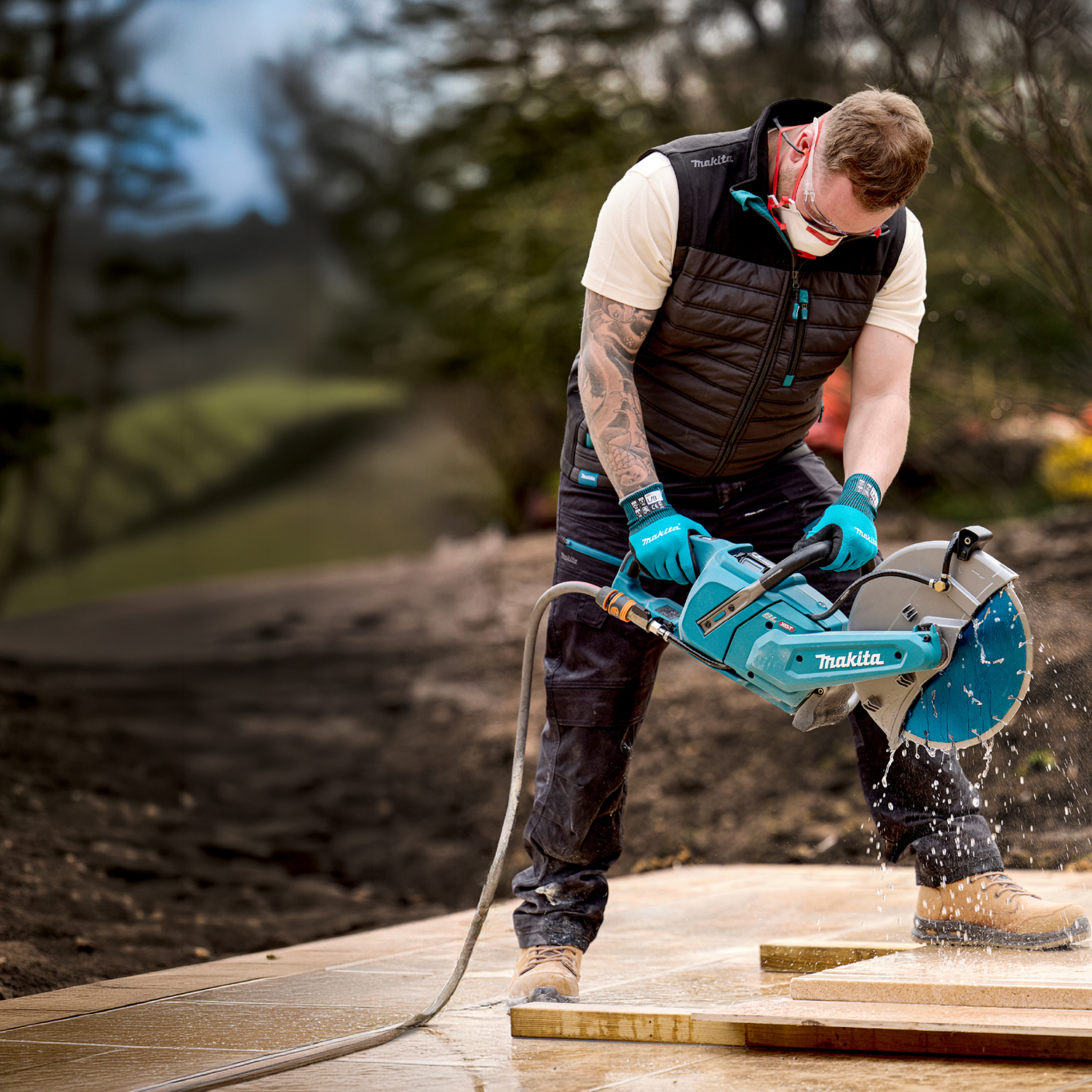 Construction worker in safety vest and gloves uses a Makita wet saw to cut wooden boards outdoors with water spraying.